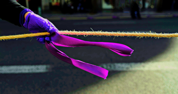 Photo of person's hand wearing purple glove and holding a purple ribbon, symbol of fight against femicide and gender violence